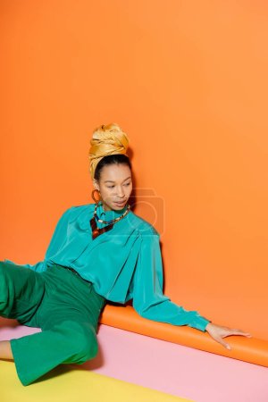Mujer afroamericana de moda en pañuelo para la cabeza y blusa azul sentada sobre fondo naranja 