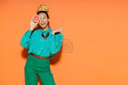 Foto de Alegre modelo afroamericano en ropa de verano con pomelo fresco sobre fondo naranja - Imagen libre de derechos
