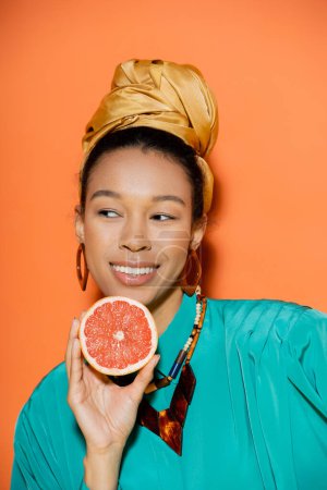 Foto de Retrato de modelo afroamericano sonriente y bien vestido sosteniendo pomelo sobre fondo naranja - Imagen libre de derechos