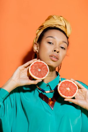 Portrait of stylish african american model with headscarf holding cut grapefruit on orange background 