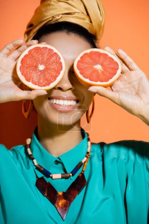 positive and stylish african american model covering face with grapefruit on orange background 