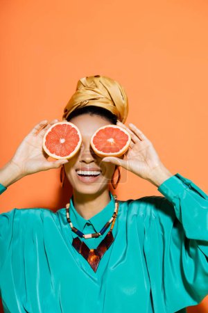 Téléchargez les photos : Femme afro-américaine positive en tenue d'été couvrant le visage avec du pamplemousse frais sur fond orange - en image libre de droit