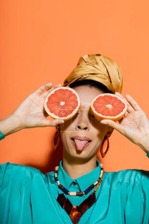 Stylish african american woman sticking out tongue and holding cut grapefruit on orange background 