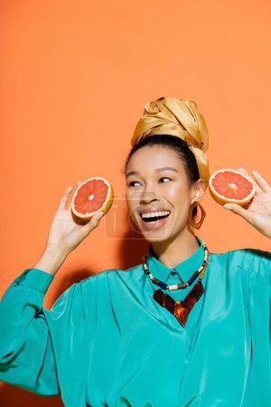 Positive african american model in bright clothes holding organic grapefruit on orange background 