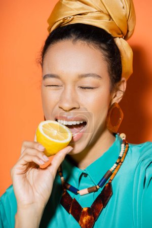 Trendige afrikanisch-amerikanische Frau beißt frische Zitrone auf orangefarbenem Hintergrund 