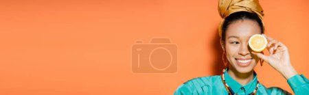 Trendy african american model smiling and holding cut lemon on orange background, banner 