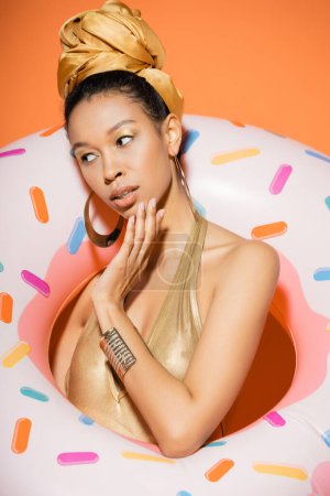 Téléchargez les photos : Portrait de modèle afro-américain tendance en maillot de bain tenant une bague de piscine sur fond orange - en image libre de droit