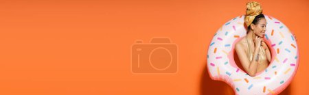 Cheerful african american model in golden headscarf posing with pool ring on orange background, banner 
