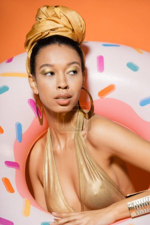 Téléchargez les photos : Portrait de modèle afro-américain à la mode en maillot de bain posant près de l'anneau de piscine sur orange - en image libre de droit