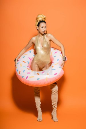Longitud completa del elegante modelo afroamericano en traje de baño con anillo de piscina sobre fondo naranja 