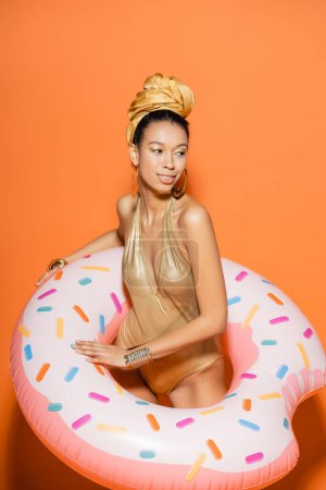 Lächelndes afrikanisch-amerikanisches Model in goldenem Badeanzug und Kopftuch mit Pool-Ring auf orangefarbenem Hintergrund 