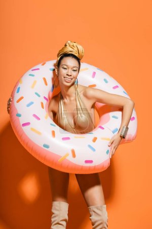 Overjoyed african american woman in swimsuit holding pool ring on orange background 
