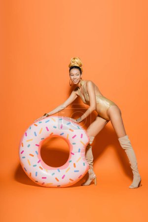 Lächelndes afrikanisch-amerikanisches Model in Kniestiefeln und Badeanzug posiert in der Nähe eines Pool-Rings auf orangefarbenem Hintergrund 