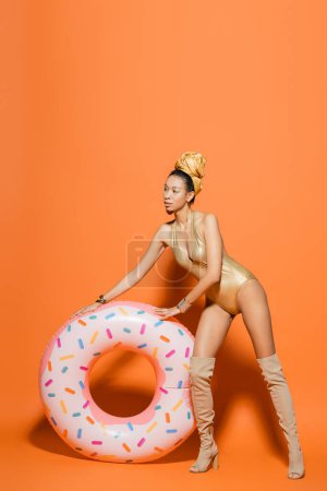 Full length of stylish african american model in golden swimsuit standing near pool ring on orange background 