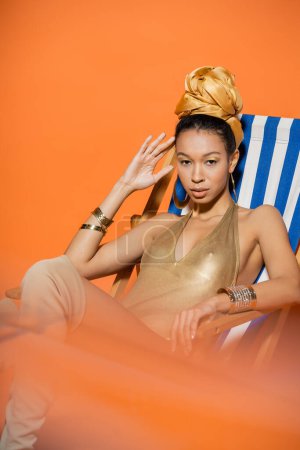 Fashionable african american model in in golden swimsuit sitting on deck chair on orange background 