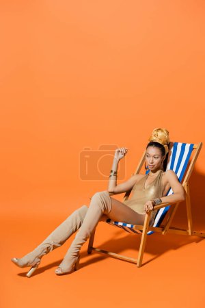 Full length of stylish african american woman in swimsuit and headscarf sitting on deck chair on orange background 