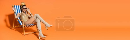 Trendy and smiling african american woman in swimwear waving hand while sitting on deck chair on orange background, banner 