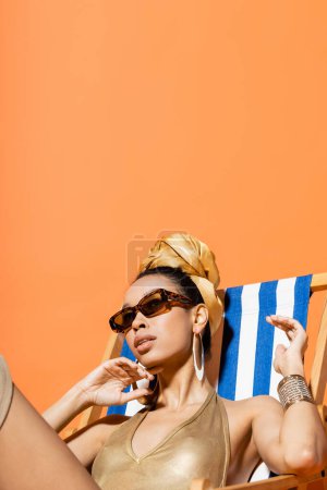 Modisches afrikanisch-amerikanisches Model in Badeanzug und Kopftuch sitzt isoliert auf einem Liegestuhl in Orange 