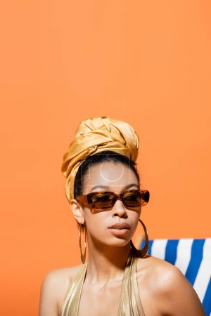 Foto de Retrato de modelo afroamericano de moda en pañuelo para la cabeza y gafas de sol mirando hacia otro lado cerca de la tumbona aislada en naranja - Imagen libre de derechos