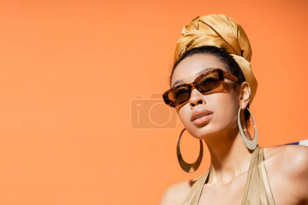 Portrait of fashionable african american model in headscarf and sunglasses isolated on orange 