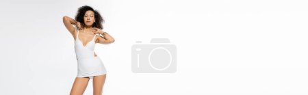 Foto de Modelo afroamericano de moda en vestido sexy posando y mirando hacia otro lado aislado en blanco, pancarta - Imagen libre de derechos