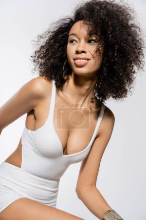 happy african american woman with curly hair posing in mini dress on grey 