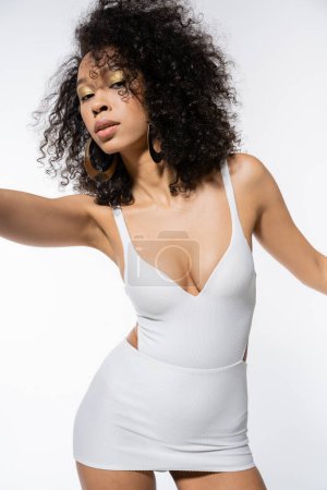 lockige Afroamerikanerin in weißem Minikleid posiert auf grauem Hintergrund 