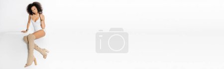 Foto de Longitud completa de rizado modelo afroamericano en mini vestido blanco y botas largas posando sobre fondo gris, bandera - Imagen libre de derechos
