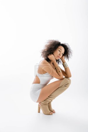 volle Länge der hübschen afrikanisch-amerikanischen Modell in Minikleid und Kniestiefel sitzt auf grauem Hintergrund 