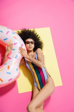 top view of pretty african american woman in swimsuit and sunglasses getting tan near inflatable ring on yellow and pink 