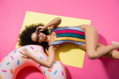 top view of african american model in swimwear adjusting sunglasses and getting tan near inflatable ring on yellow and pink  Poster #648703030