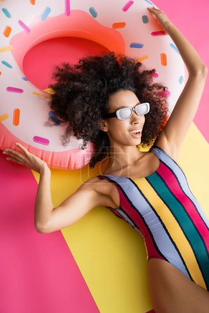 positives afrikanisch-amerikanisches Model in gestreifter Badebekleidung und Sonnenbrille, die sich in der Nähe eines aufblasbaren Rings in gelb und rosa bräunt 