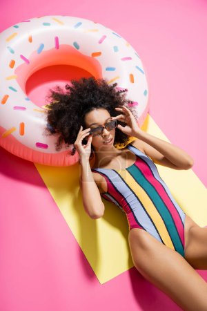 top view of african american woman in striped swimwear adjusting sunglasses and getting tan near inflatable ring on yellow and pink 