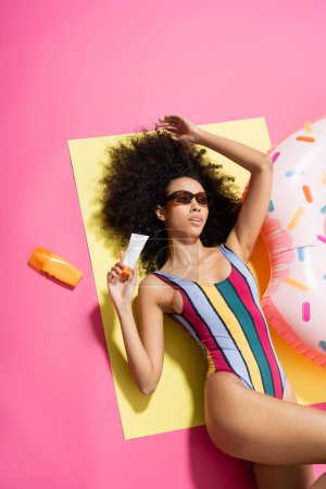 top view of african american woman in striped swimwear and trendy sunglasses holding sunscreen near inflatable ring on pink