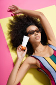 top view of pretty african american woman in trendy sunglasses and striped swimwear holding sunscreen while getting tan on pink puzzle #648703240