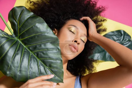 top view of curly african american woman with closed eyes getting tan near green palm leaves on pink 