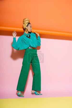 full length of happy african american woman in stylish outfit smiling while holding rolled orange wallpaper on pink background 