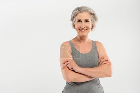 mujer feliz y mayor en ropa deportiva de pie con los brazos cruzados aislados en gris
