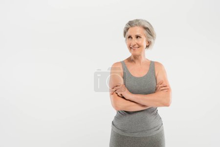 fröhliche und ältere Frau in Sportkleidung mit verschränkten Armen, vereinzelt auf grau