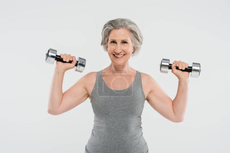 excité femme âgée avec les cheveux gris exercice avec haltères isolé sur gris 
