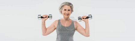 femme âgée ravie avec des cheveux gris exercice avec haltères isolé sur gris, bannière  