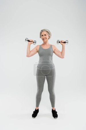 full length of happy senior woman in tight sportswear exercising with dumbbells on grey 