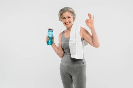 happy senior woman with white towel on shoulder holding sports bottle with water and showing ok isolated on grey 
