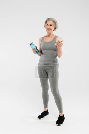 Foto de Longitud completa de la mujer mayor feliz en ropa deportiva celebración de botella de deportes con agua y teléfono inteligente en gris - Imagen libre de derechos