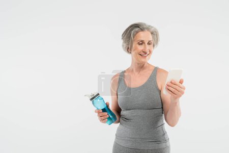 glückliche Seniorin in Sportkleidung, die Sportflasche mit Wasser in der Hand und Smartphone isoliert auf grau 