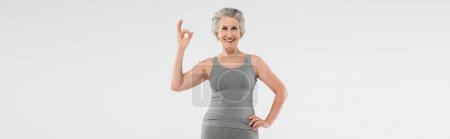 Foto de Mujer alegre y jubilada en ropa deportiva posando con la mano en la cadera y mostrando bien aislado en gris, bandera - Imagen libre de derechos