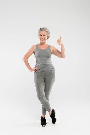 Foto de Longitud completa de mujer alegre y jubilada en ropa deportiva posando con la mano en la cadera y mostrando como en gris - Imagen libre de derechos