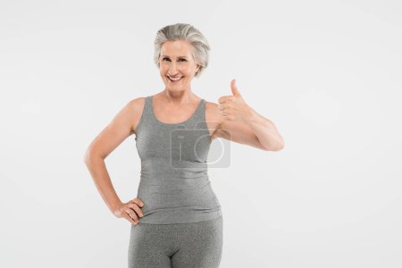 fröhliche Rentnerin in Sportbekleidung posiert mit der Hand an der Hüfte und zeigt sich wie vereinzelt auf grau 