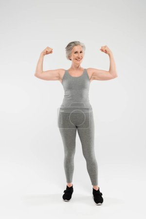 lebensfrohe Rentnerin in Sportbekleidung, die Muskeln auf grau zeigt 