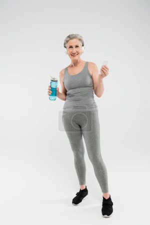 full length of happy senior woman listening music in headphones while holding sports bottle and smartphone on grey 
